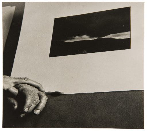 A weathered hand rests on a dark table beside a small mounted landscape photograph
